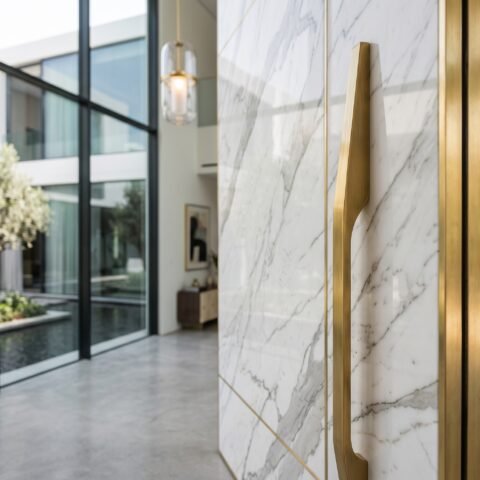Double heighted white marble pivot door with gray veining, gold trim and vertical handle in a luxury modern interior