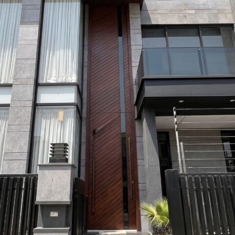 Massive dark wood double heighted pivot door with diagonal patterns and a glass slit on a modern grey stone facade.