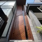 Massive dark wood double heighted pivot door with diagonal patterns and a glass slit on a modern grey stone facade.