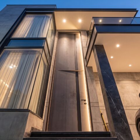 A towering matte grey double heighted pivot door with a vertical LED light strip installed in a luxury modern villa by Tarkhan Door.