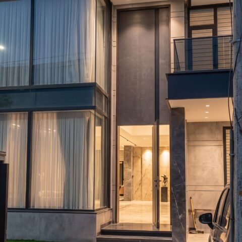 A towering matte grey double heighted pivot door with a vertical LED light strip installed in a luxury modern villa by Tarkhan Door.