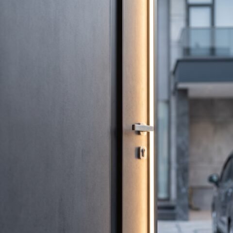 A towering matte grey double heighted pivot door with a vertical LED light strip installed in a luxury modern villa by Tarkhan Door.