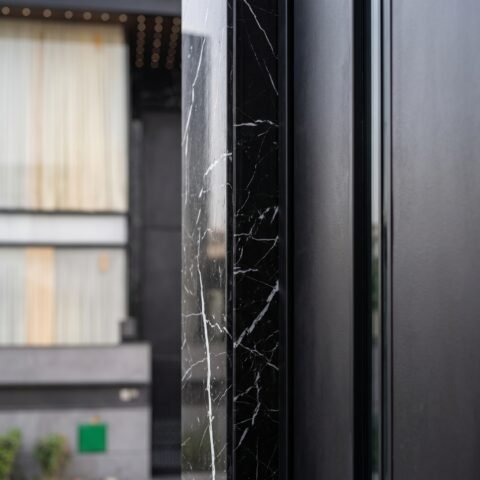 A massive matte black double heighted pivot door framed by black veined marble on a luxury modern residence by Tarkhan Door.